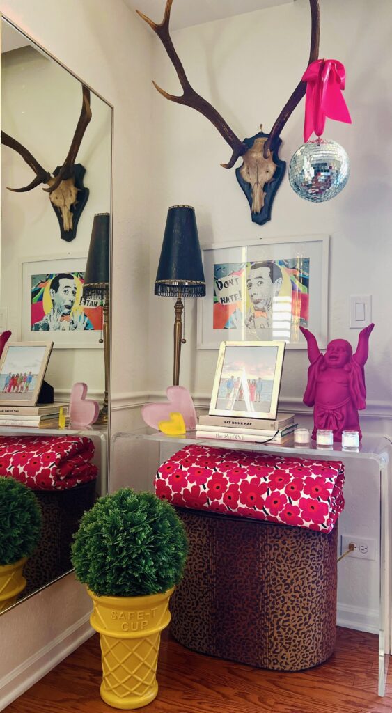 Eclectic room features a mounted antler with disco ball, colorful pop art, pink Buddha statue, mirrored reflection, and decor on a leopard print ottoman.