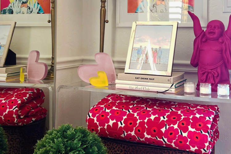 A decorative table with a pink laughing Buddha statue, framed family photo, and candles. Stacks of books, pink and yellow heart decorations, and a red floral blanket create a cozy, playful ambiance.