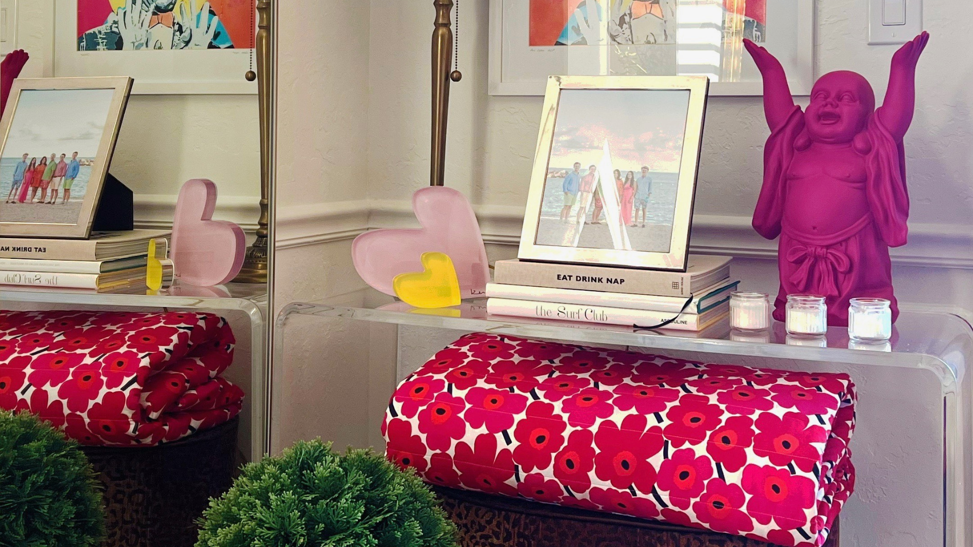 A decorative table with a pink laughing Buddha statue, framed family photo, and candles. Stacks of books, pink and yellow heart decorations, and a red floral blanket create a cozy, playful ambiance.